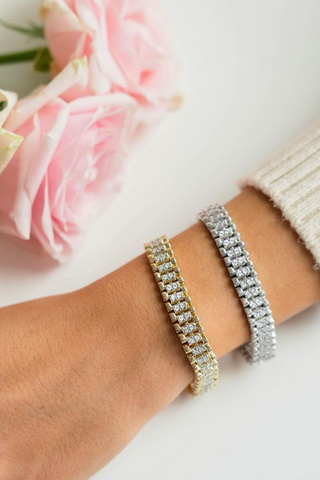 Two hands wearing gold and silver bracelets with pink roses in the background